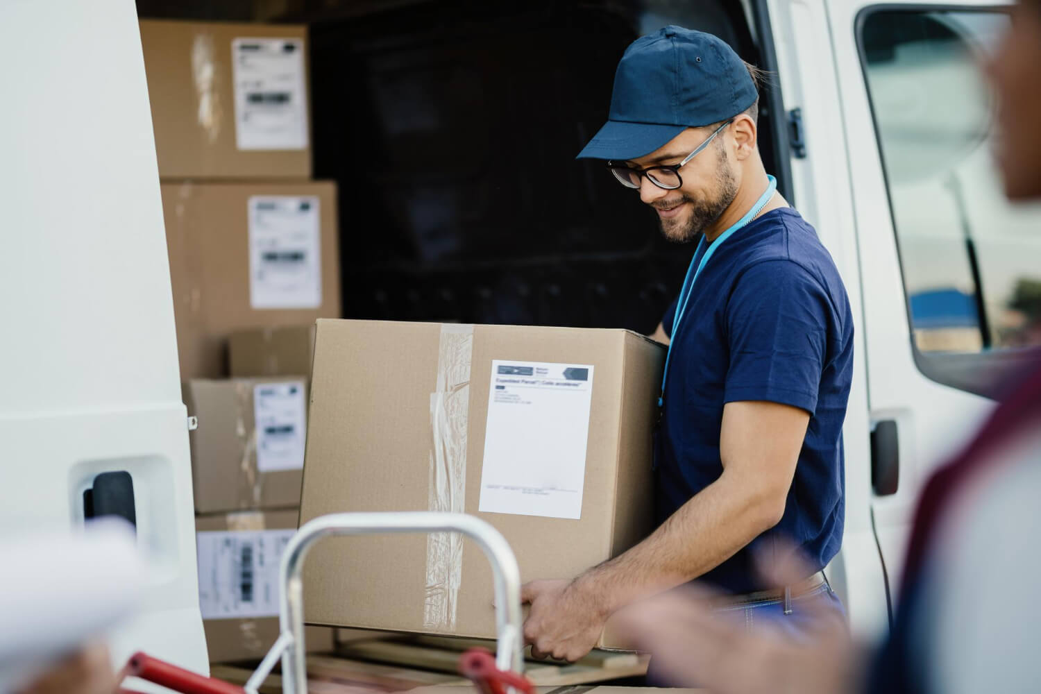 Delivery worker in navy blue shirt and cap carrying a large cardboard box near a van loaded with packages.